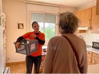 Une femme en chemise orange tient un panier en plastique tout en discutant avec une femme plus âgée aux cheveux gris dans une cuisine avec des armoires en bois et une fenêtre.