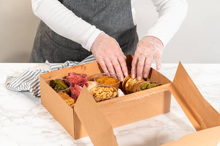 Une personne portant des gants et un tablier gris emballe une boîte en carton contenant un assortiment d'en-cas (crackers, noix, charcuterie et cornichons) sur un comptoir en marbre blanc.